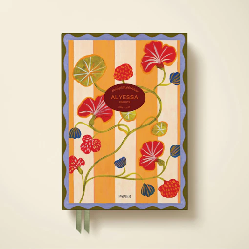 The photo shows the cover of a planner from Papier. The background of the planner features thick yellow and cream vertical stripes with a green and blue squiggly border. A pattern of red and blue flowers and leaves overlays the background, and a maroon oval contains the name and information of the planner's owner in orange letters.